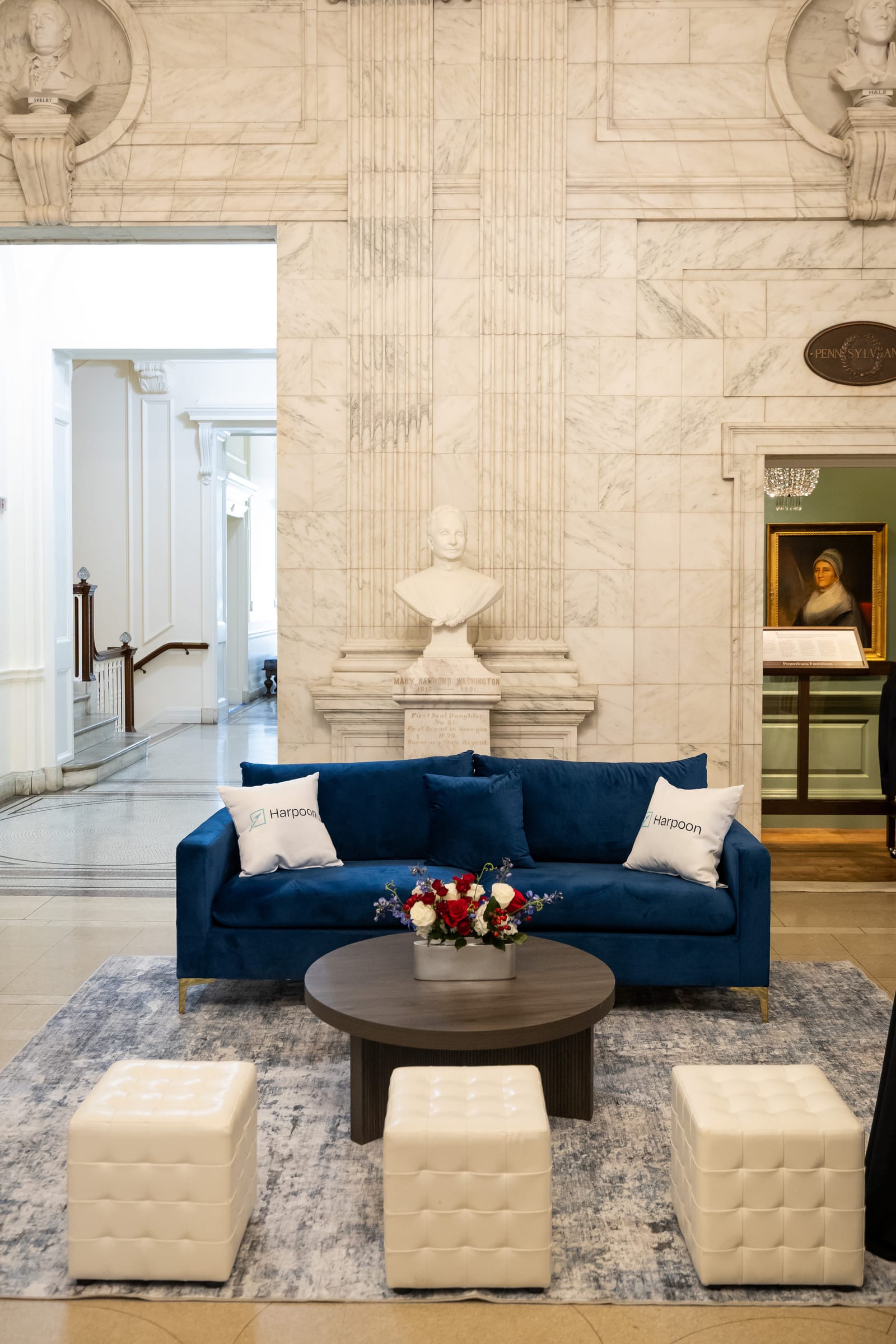Freedom Tech Summit lounge setting Summit lounge with blue velvet sofa, Harpoon pillows, tufted ottomans, coffee table, and rug beneath a classical bust in the marble hall