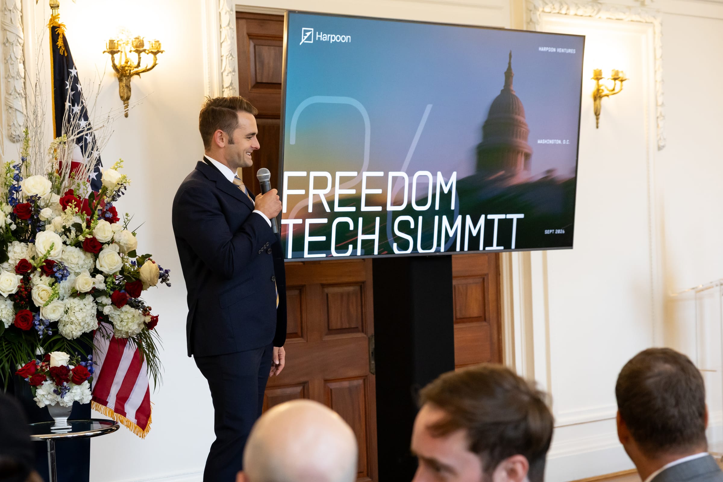Freedom Tech Summit opening remarks Speaker delivering opening remarks beside a Freedom Tech Summit screen and patriotic floral arrangement