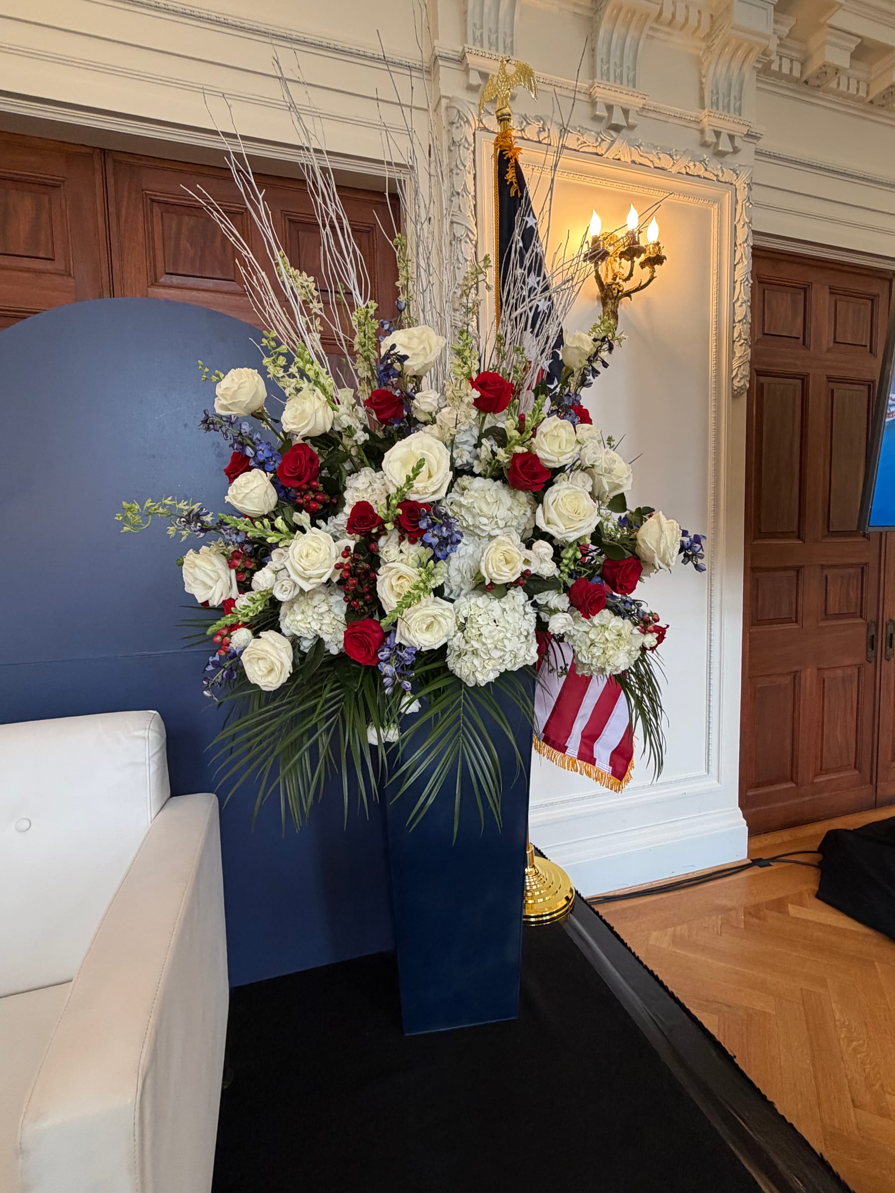 Freedom Tech Summit stage floral design Red, white, and blue floral arrangement with roses, hydrangeas, and white branches in a navy pedestal beside the stage flag