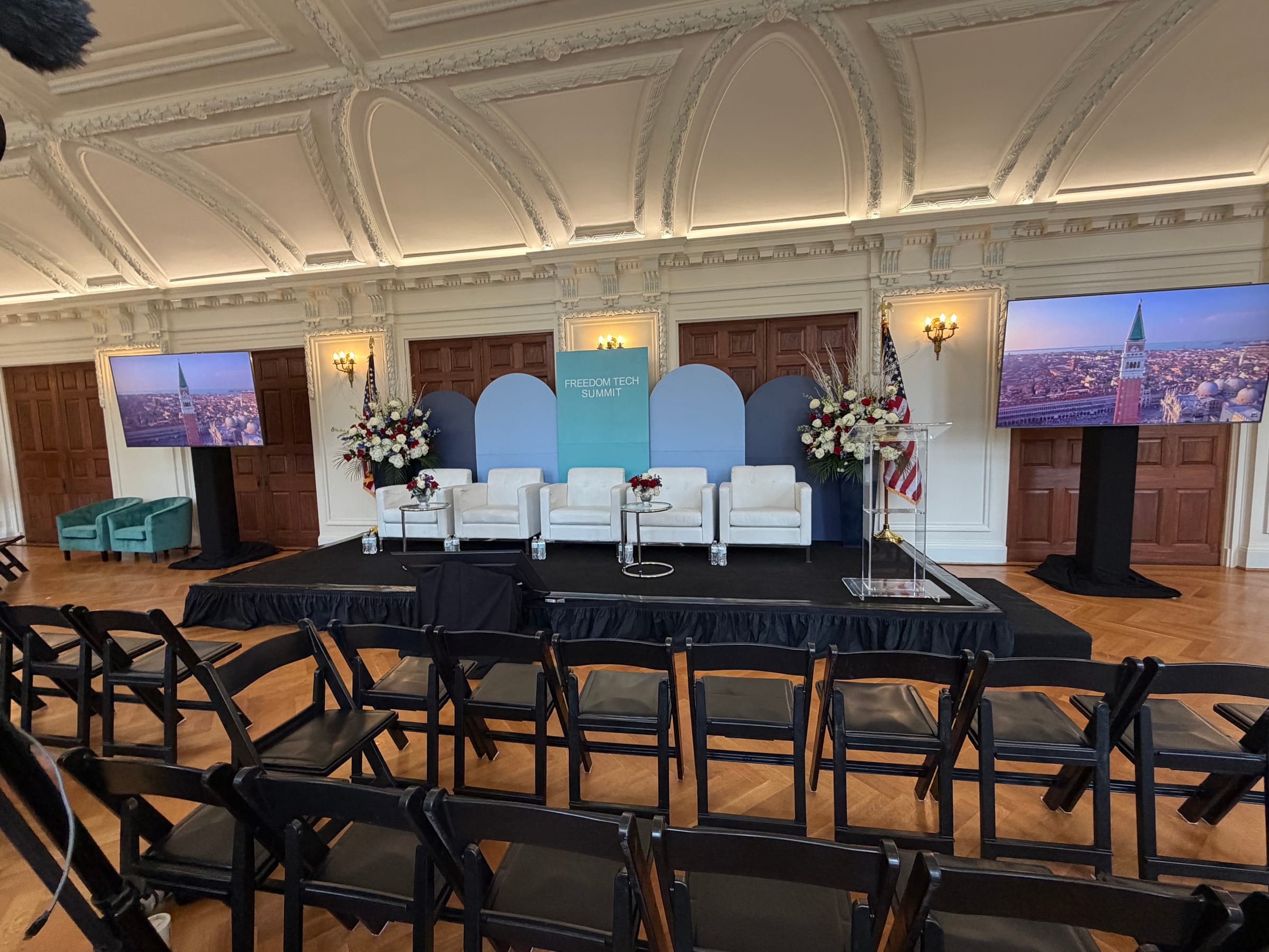Freedom Tech Summit stage and audience seating Wide view of the Freedom Tech Summit stage with arched scenic backdrop, florals, flanking screens, and audience seating